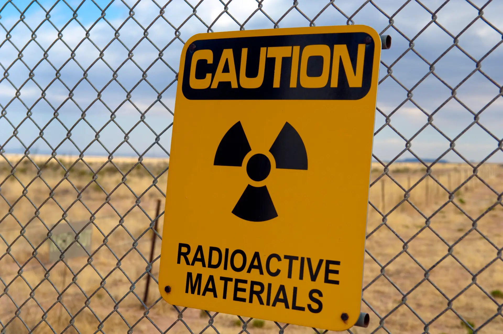 Drummond Carpenter nuclear solutions precision and defensibility approach illustrated by a caution radioactive materials warning sign with radiation hazard symbol mounted on a chain-link security fence at a remote nuclear site, representing the firm's rigorous standards for radiological safety compliance, defensible data collection, precise contamination assessment, and regulatory-grade nuclear site documentation.
