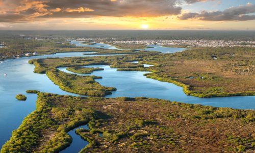 Drummond Carpenter environment solutions regulatory fluency approach illustrated by a sweeping panoramic aerial view of a protected coastal river delta at sunset, with intricate mangrove-lined waterways, wetland ecosystems, and residential communities along the shoreline, representing the firm's deep expertise in environmental regulatory compliance, wetland permitting, waterway jurisdictional determinations, and navigating complex federal and state environmental regulations.