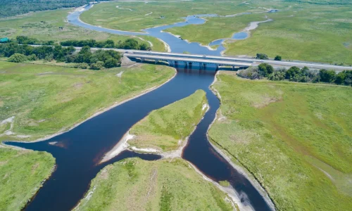 Drummond Carpenter water solutions systems perspective illustrated by an aerial view of a branching river watershed flowing through expansive wetland floodplains and beneath a highway bridge, representing the firm's holistic approach to watershed management, hydrological systems analysis, stormwater planning, and integrated water infrastructure engineering.