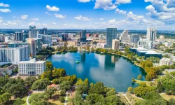 Drummond Carpenter state and local government and education services illustrated by a vibrant aerial view of a thriving downtown urban center surrounding a large municipal lake with parks, mixed-use development, and modern high-rise buildings under a bright blue sky, representing the firm's expertise supporting municipalities, regional agencies, water management districts, universities, and local governments with infrastructure engineering, water systems planning, and community development services.